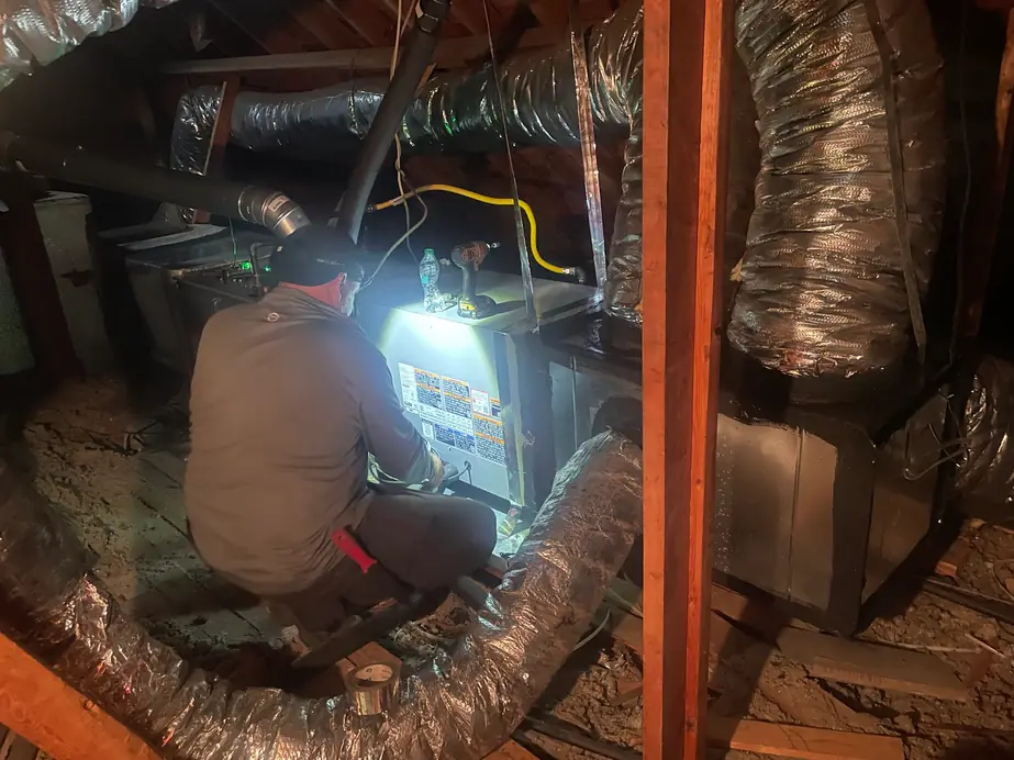 Technician wearing a headlamp working at an air handler surrounded by attic ductwork